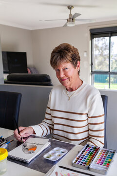happy senior woman working on watercolour painting in living room at home