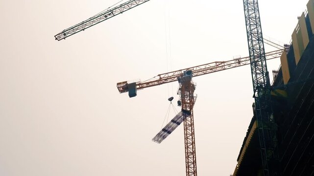 Tower crane lifting bundled rebar over high rise construction site under pale sky, low angle view emphasizing scale and silhouetted scaffolding