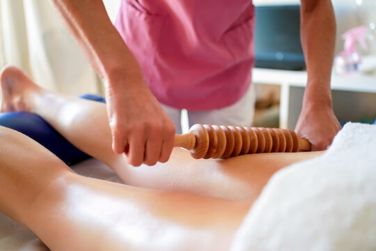 Therapist using wooden massage roller across client back during spa treatment in bright wellness room with soft warm light and calm clinical atmosphere, with massage, therapist, roller, wooden detail