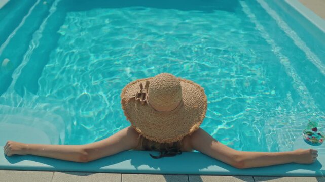 Woman wearing straw hat relaxing in blue water of swimming pool during summer vacation at sunny day.