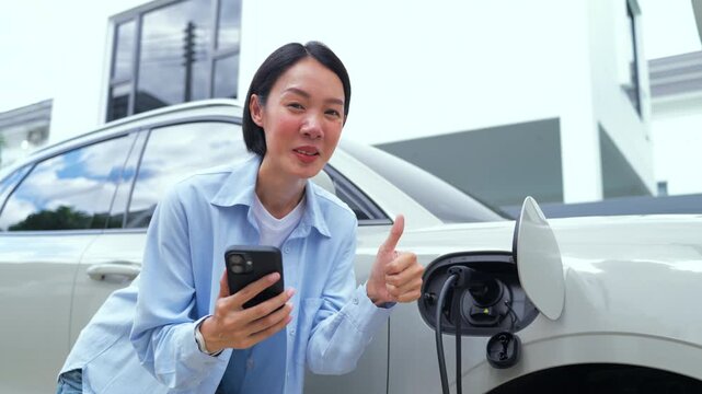 Smiling Asian woman showing a thumbs up while holding a smartphone and charging her electric car at home. Smart EV home charging and clean energy concept.