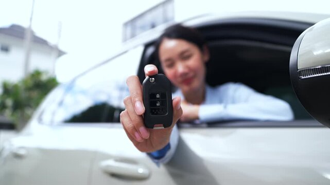 Close-up of a smiling Asian woman holding and showing a car key fob out of her electric vehicle window at home. New car owner and sustainable lifestyle concept.