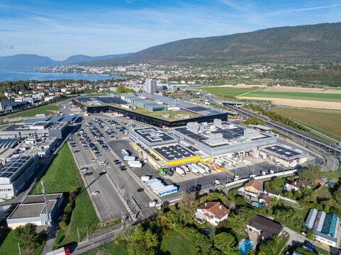 Aerial view of Marin Centre shopping complex, featuring large flat rooftops with solar panels, loading docks with trucks, and parking lots in Latena, Neuchatel, Switzerland.