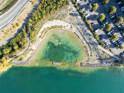 Aerial view of a small bay with turquoise water, rocky shoreline, adjacent parking lot, and surrounding greenery in Neuenburg, Neuenburg, Switzerland.