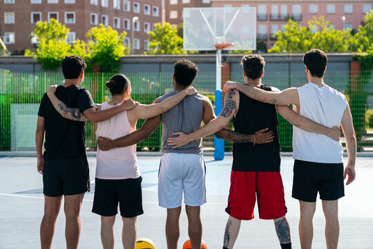 Group of diverse friends with arms around each other, standing on a sunny basketball court, showing teamwork and unity