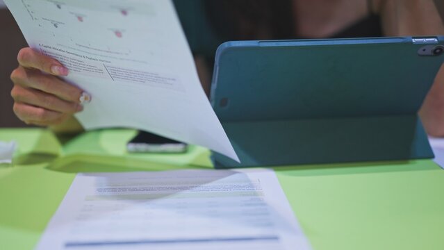 Analyst reviews printed report sheets beside digital tablet upon work desk, examining corporate capital allocation metrics, tracking investment budget forecasts, evaluating market trends.