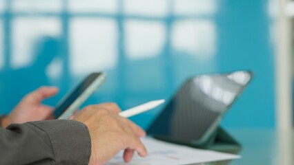 Businessman holding stylus pen mobile phone over financial report papers beside digital tablet on desk in bright corporate office setting. © Mdisk
