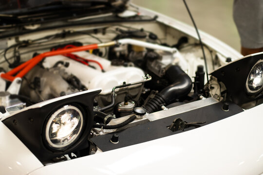 Open white car hood showing engine bay with red strut bar, black intake tubing, exposed headlights, and mechanical components in an automotive workshop