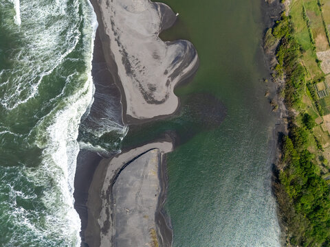 Aerial view of Depok Beach where the river meets the ocean with dark sand banks and lush green vegetation in Special Region of Yogyakarta, Indonesia.