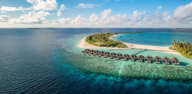 Aerial panoramic overwater villas turquoise tropical island ocean sky sand coral reef resort paradise. Peaceful summer nature seascape scene. Luxury honeymoon drone travel scenic coastal vacation