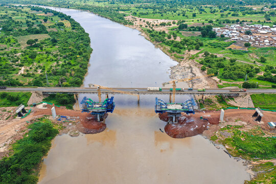 Aerial view of the construction site of the new Yapei Bridge over the White Volta River, featuring heavy machinery and cranes alongside the existing bridge, Yapei, Savannah Region, Ghana.