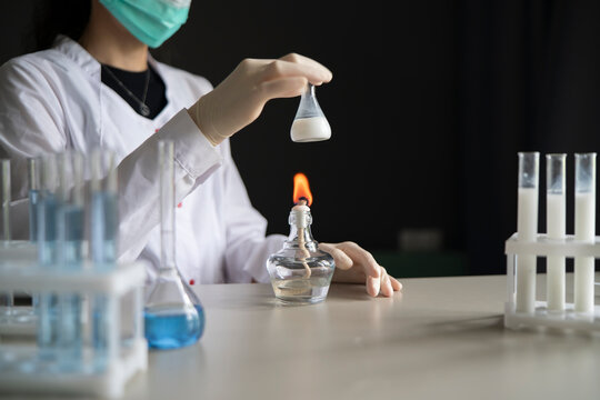 Laboratory Worker Holding Small Flask of White Liquid Over Burning Wick Lamp With Test Tube Racks Nearby