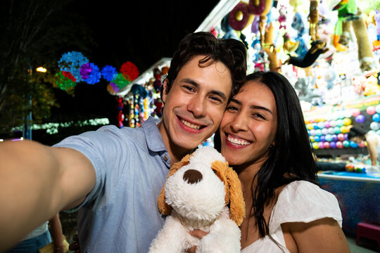 Smiling young couple taking a selfie with a plush toy dog at a nighttime carnival