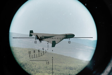 A military drone flies over green hills and is tracked in an optical surveillance scope. © Ernest
