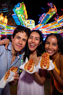 Diverse friends enjoying hot dogs, smiling, having fun at amusement park