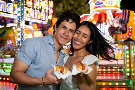Smiling young man and woman holding hot dogs at a vibrant night carnival