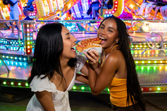 Two cheerful young women sharing a hot dog at a vibrant night fair