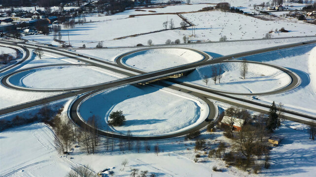 Aerial view of a snow-covered cloverleaf highway interchange surrounded by winter fields and trees in Latvia.