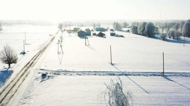 Aerial view of a snow-covered rural landscape with scattered farm buildings, frost-covered trees, and a quiet road under bright winter sunlight in Latvia.