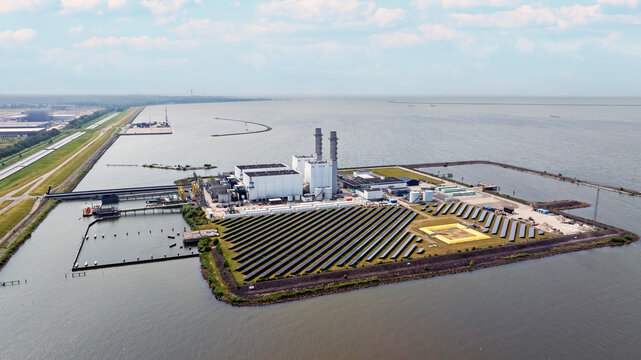 Aerial view of the Maxima power plant with its solar panel field, industrial buildings, and chimneys situated on a dike along the water in Lelystad, Flevoland, Netherlands.