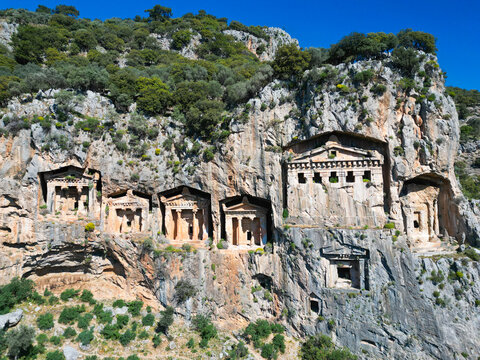 Aerial view of the ancient Lycian rock tombs carved into the cliff face surrounded by rugged rock formations and green vegetation in Dalyan, Mugla, Turkiye.