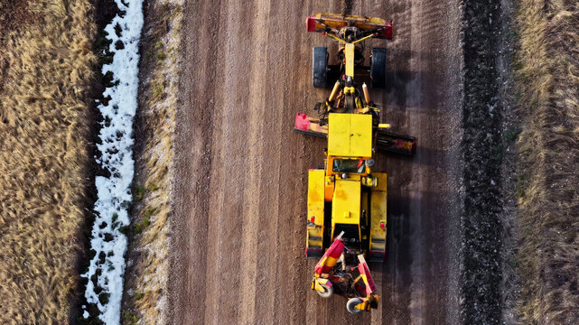 Aerial view of a yellow motor grader leveling a dirt road with patches of snow on the roadside Latvia.