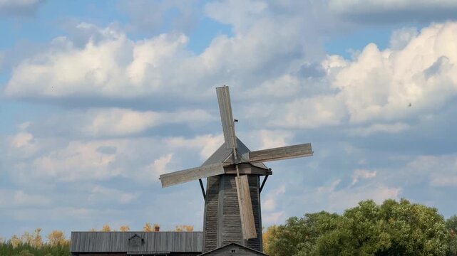 Against the backdrop of a azure sky with fluffy clouds, an old wooden windmill rises, surrounded by green trees.
