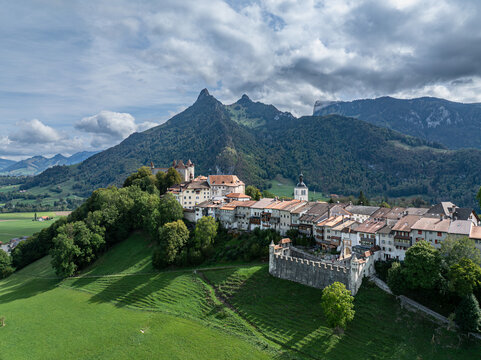 Aerial view of Chateau de Gruyeres perched on a hill above the historic town, surrounded by green meadows, forests, and Prealps peaks under a cloudy sky, Gruyeres, Fribourg, Switzerland.