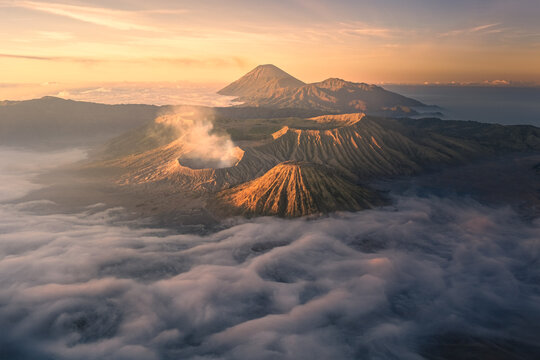 Aerial view of Mount Bromo with its volcanic crater emitting smoke, surrounded by a sea of clouds at sunrise in Tosari, East Java, Indonesia.