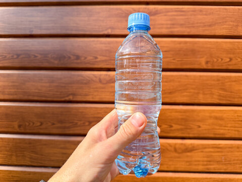 Human hand holding a clear plastic bottle of pure drinking water against a brown wooden wall background outdoors. Clean mineral water bottle template for healthy lifestyle and hydration theme.