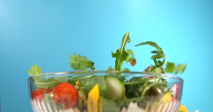 Fresh vegetable salad ingredients falling into glass bowl in slow motion on bright blue background