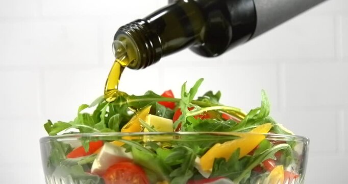 Olive oil pouring from bottle onto fresh vegetable salad with arugula tomato pepper and white cheese