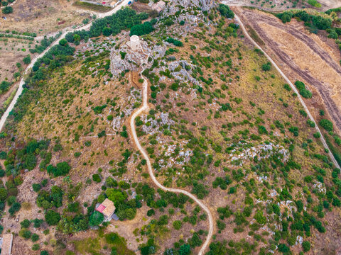 Aerial view of a historic stone watchtower atop a rugged hill with a winding dirt path surrounded by dry Mediterranean vegetation in Palermo, Sicily, Italy.
