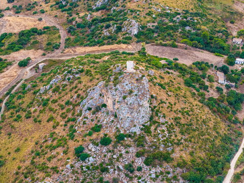 Aerial view of a rocky hill topped with a historic stone watchtower, surrounded by Mediterranean scrub, dry grass, and paths in the rural landscape of Palermo, Sicily, Italy.