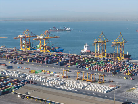 Aerial view of the Port of Thessaloniki with rows of colorful shipping containers, large yellow gantry cranes, and cargo ships docked in the Aegean Sea, Thessaloniki, Greece.