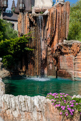 Artificial rock waterfall cascading over stone formations into turquoise pool surrounded by pink flowers, luxury resort landscape closeup with lush green foliage, high quality photo © Татьяна Швец