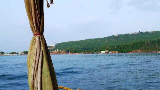 A tied-back boat curtain frames Lake Pichola, a green hillside and the boat's foaming wake in Udaipur.