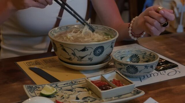 Diner lifts noodle strands with chopsticks from patterned ceramic soup bowl on warm wooden table, with clear broth, bean sprouts, herbs, chili and lime nearby in close view