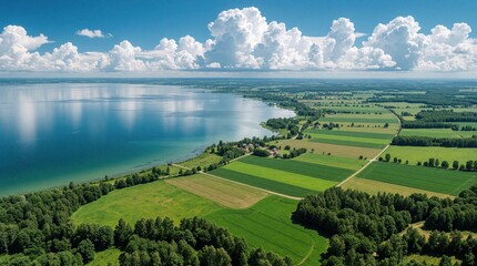Fototapeta premium Aerial view of calm blue lake, green farmland. Large white clouds float in bright sky over rural countryside. Fields, forests meet water shore. Scenic landscape shows peaceful nature scenery with