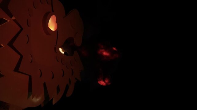 Dragon head sculpture silhouette breathing bright fireball into night darkness, with drifting smoke plume and glowing embers around carved mouth and eye cutouts