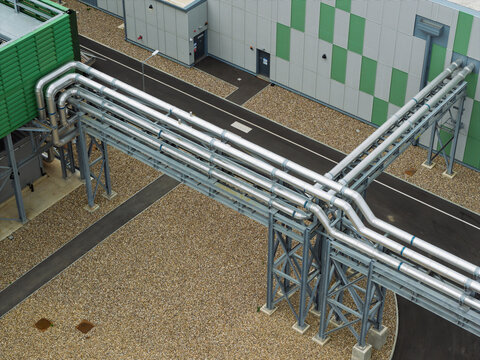 Aerial view of a modern data center facility featuring complex industrial piping systems, metal support structures, and gravel ground surfaces in the United Kingdom.