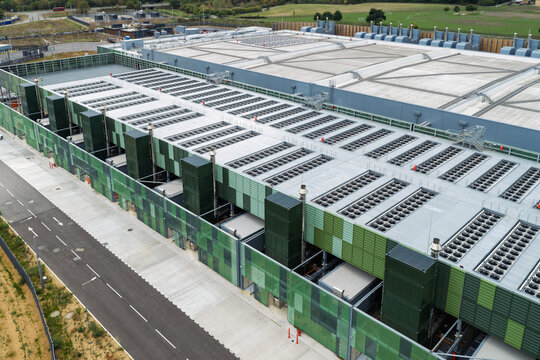 Aerial view of a modern data center facility featuring green and grey modular cladding, extensive rooftop cooling infrastructure, and a paved access road in the United Kingdom.