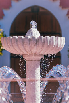 Close view of a scalloped marble fountain with sparkling droplets and curved water jets set against a softly blurred arched doorway.