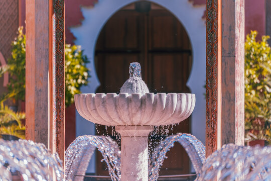 A white fountain and curving water jets align between decorated pavilion columns before a dark arched wooden door in warm sunlight.