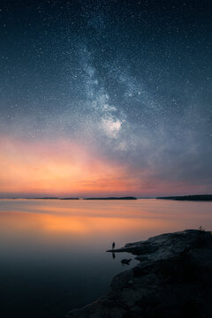 Silhouette of a lone person standing on a rocky shore under the Milky Way and sunset sky over a calm lake