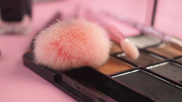 Close up of pink fluffy makeup brush and eyeshadow palette on pink surface.