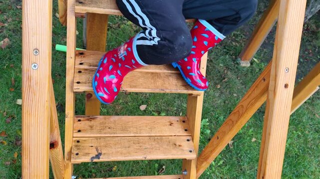 Child wearing red rain boots and black pants climbing wooden outdoor steps in a garden.
