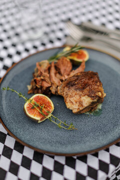 Cooked lamb pieces with fresh figs, rosemary, and thyme on a blue ceramic plate, set on a black-and-white checkered tablecloth with cutlery