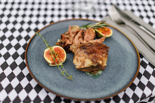 Cooked lamb pieces with fresh figs, rosemary, and thyme on a blue ceramic plate, set on a black-and-white checkered tablecloth with cutlery