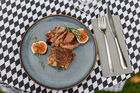 Sliced cooked lamb arranged on a ceramic plate with fresh figs, rosemary, and thyme, served with cutlery on a black-and-white checkered tablecloth